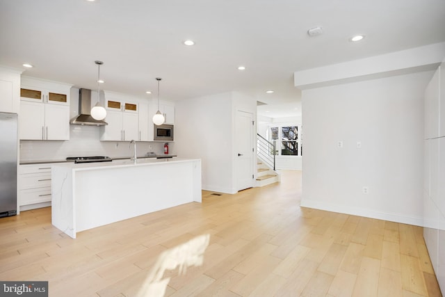 kitchen featuring light hardwood / wood-style flooring, wall chimney exhaust hood, an island with sink, stainless steel appliances, and pendant lighting