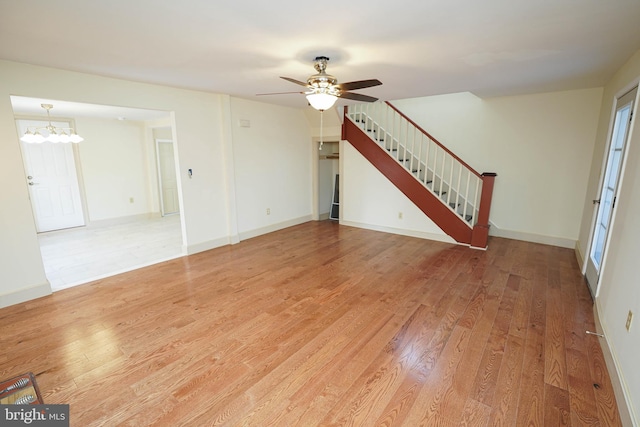 unfurnished living room with stairway, light wood-style flooring, ceiling fan with notable chandelier, and baseboards