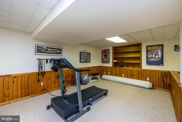 exercise area featuring light colored carpet, a drop ceiling, built in features, and wood walls