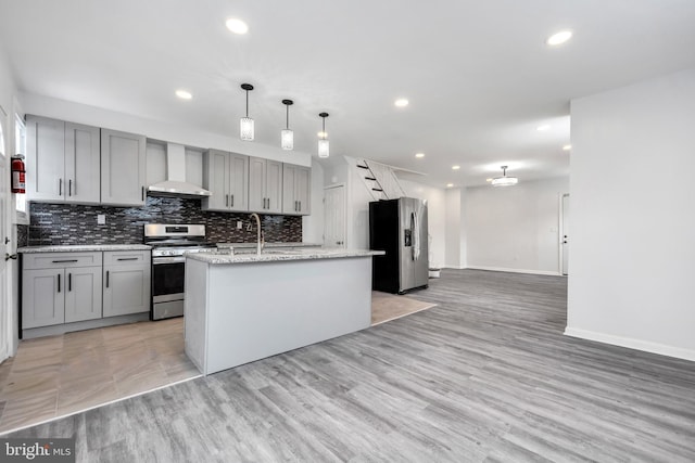 kitchen featuring wall chimney exhaust hood, gray cabinetry, a center island with sink, pendant lighting, and stainless steel appliances