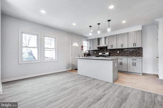 kitchen featuring pendant lighting, wall chimney range hood, gray cabinets, a kitchen island with sink, and stainless steel range with electric stovetop