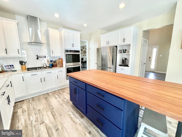 kitchen with appliances with stainless steel finishes, wood counters, blue cabinets, white cabinets, and wall chimney range hood