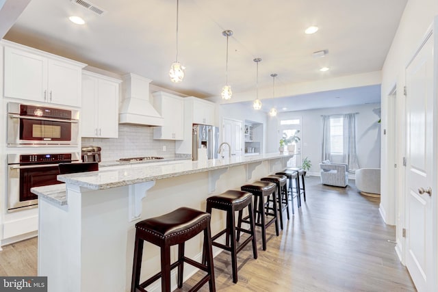 kitchen featuring premium range hood, white cabinetry, decorative light fixtures, stainless steel appliances, and a kitchen island with sink