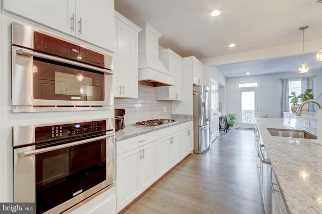 kitchen featuring appliances with stainless steel finishes, white cabinetry, sink, light stone counters, and custom range hood