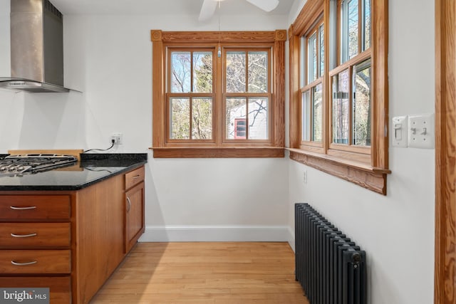 kitchen featuring light hardwood / wood-style flooring, radiator heating unit, wall chimney exhaust hood, stainless steel gas stovetop, and dark stone countertops