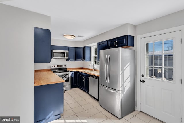 kitchen with sink, light tile patterned flooring, appliances with stainless steel finishes, blue cabinets, and wood counters