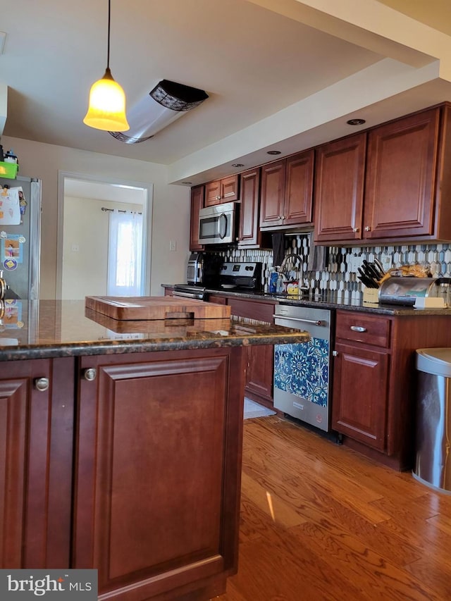 kitchen featuring appliances with stainless steel finishes, pendant lighting, backsplash, dark stone counters, and light hardwood / wood-style flooring