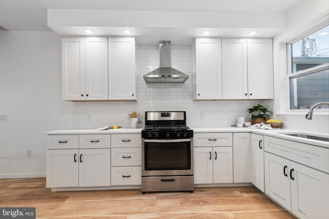 kitchen featuring wall chimney range hood, sink, white cabinetry, light hardwood / wood-style floors, and stainless steel range with gas cooktop