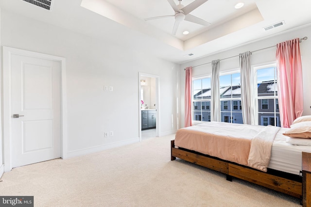 bedroom with ceiling fan, light colored carpet, a tray ceiling, and ensuite bath