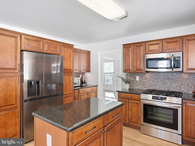 kitchen with appliances with stainless steel finishes, backsplash, light wood-type flooring, dark stone countertops, and a kitchen island