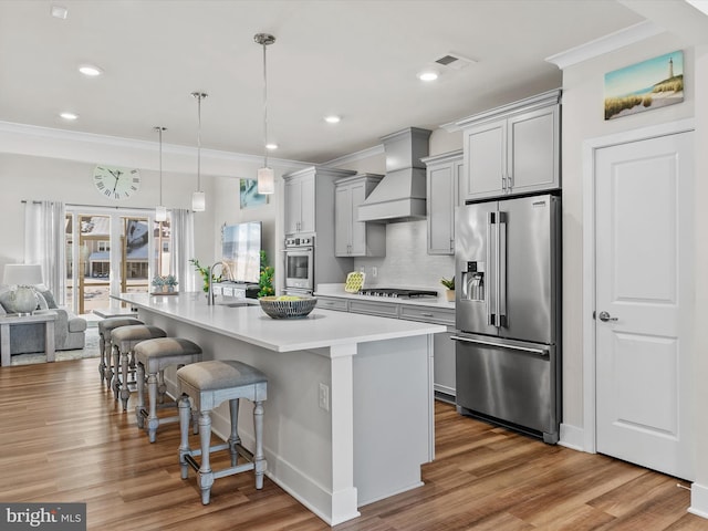 kitchen featuring appliances with stainless steel finishes, premium range hood, gray cabinetry, an island with sink, and a breakfast bar area