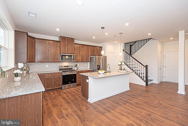 kitchen with tasteful backsplash, decorative light fixtures, a center island with sink, appliances with stainless steel finishes, and light stone countertops