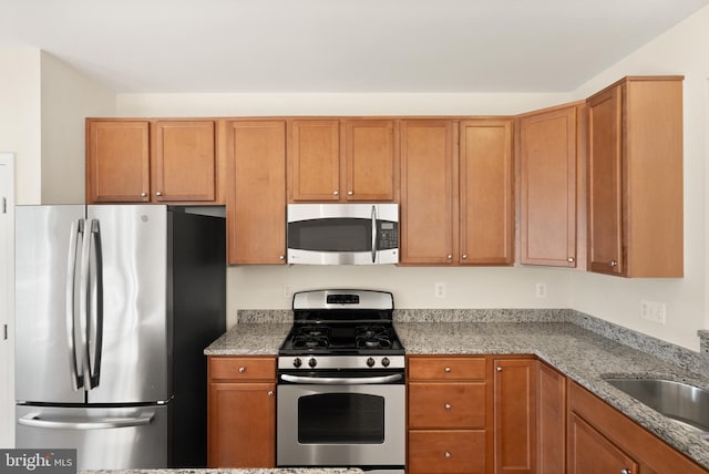 kitchen featuring brown cabinetry, appliances with stainless steel finishes, light stone counters, and a sink