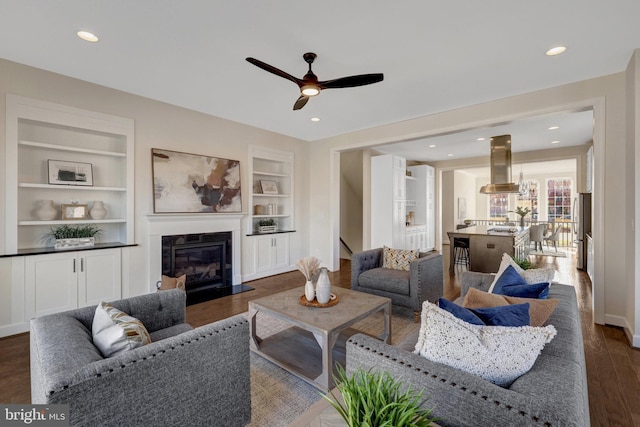 living area with baseboards, dark wood finished floors, a glass covered fireplace, built in shelves, and recessed lighting