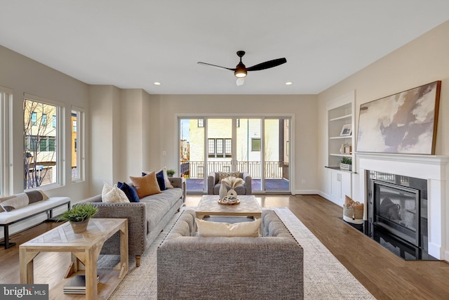 living room featuring baseboards, a glass covered fireplace, wood finished floors, built in shelves, and recessed lighting