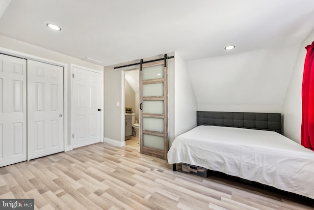 bedroom featuring a barn door, vaulted ceiling, connected bathroom, and light hardwood / wood-style floors