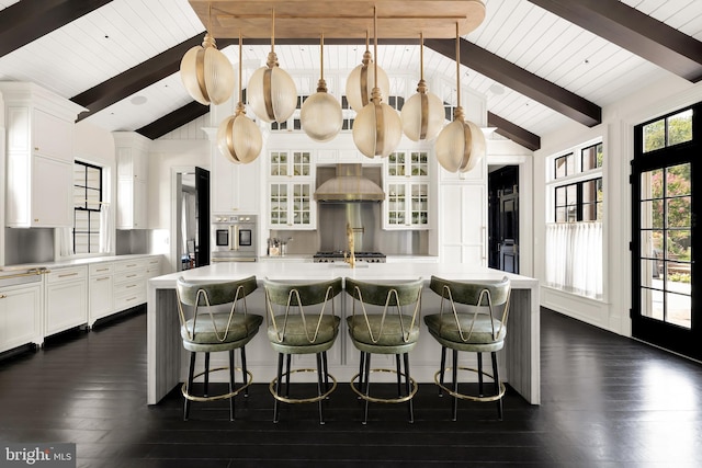 kitchen with a center island with sink, stainless steel oven, hanging light fixtures, white cabinets, and wall chimney exhaust hood