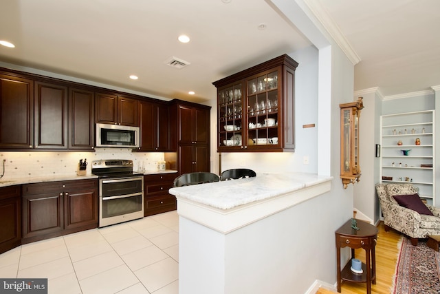 kitchen with dark brown cabinetry, crown molding, tasteful backsplash, light tile patterned floors, and appliances with stainless steel finishes