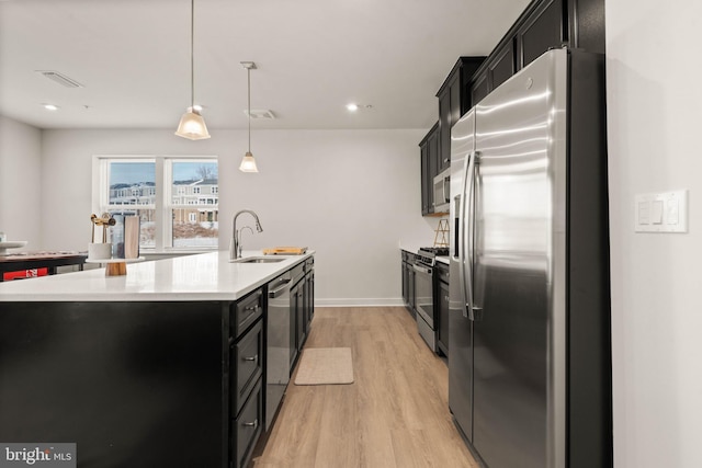 kitchen with sink, decorative light fixtures, light wood-type flooring, appliances with stainless steel finishes, and a kitchen island with sink