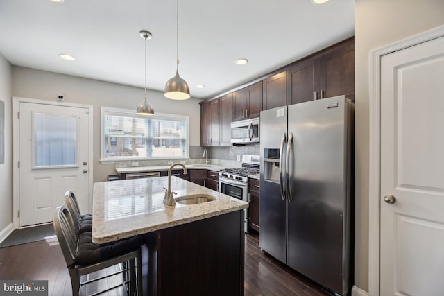 kitchen featuring an island with sink, appliances with stainless steel finishes, dark brown cabinets, light stone countertops, and sink