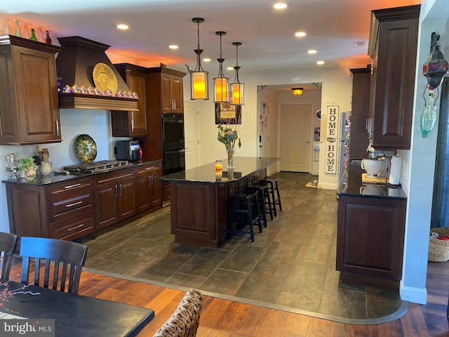 kitchen with stainless steel gas stovetop, black double oven, custom range hood, hanging light fixtures, and a kitchen island