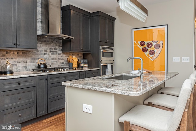 kitchen featuring a center island with sink, a breakfast bar area, appliances with stainless steel finishes, tasteful backsplash, and wall chimney range hood