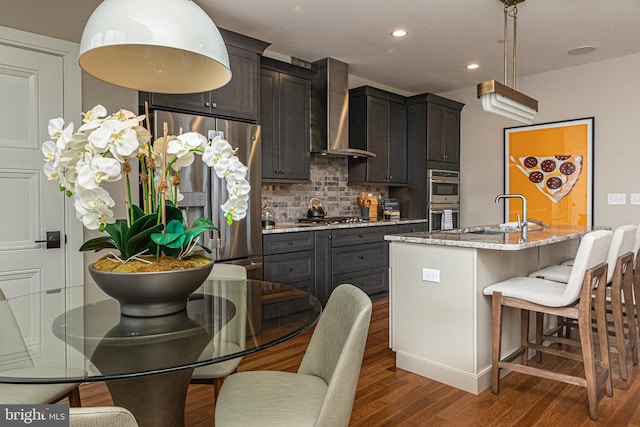 kitchen featuring a center island with sink, stainless steel appliances, backsplash, wall chimney exhaust hood, and sink