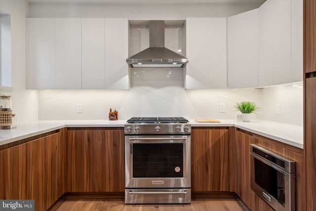 kitchen with wall chimney range hood, light hardwood / wood-style floors, white cabinetry, and appliances with stainless steel finishes