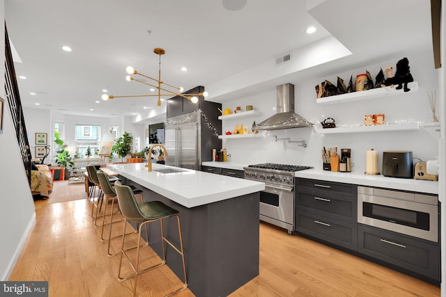 kitchen with wall chimney range hood, light hardwood / wood-style floors, sink, high end appliances, and an island with sink