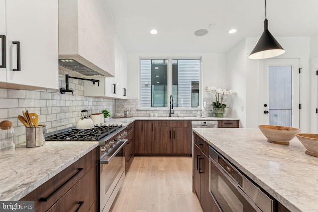 kitchen featuring light stone countertops, white cabinets, appliances with stainless steel finishes, decorative light fixtures, and wall chimney range hood