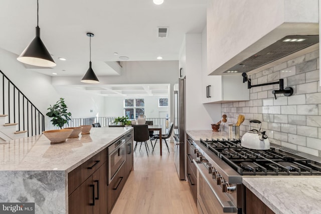 kitchen with hanging light fixtures, light stone countertops, wall chimney exhaust hood, high quality appliances, and light hardwood / wood-style flooring