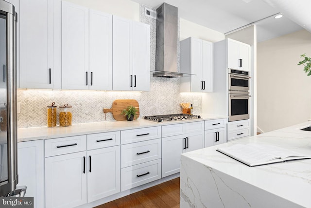 kitchen with light stone countertops, appliances with stainless steel finishes, white cabinets, wall chimney range hood, and decorative backsplash