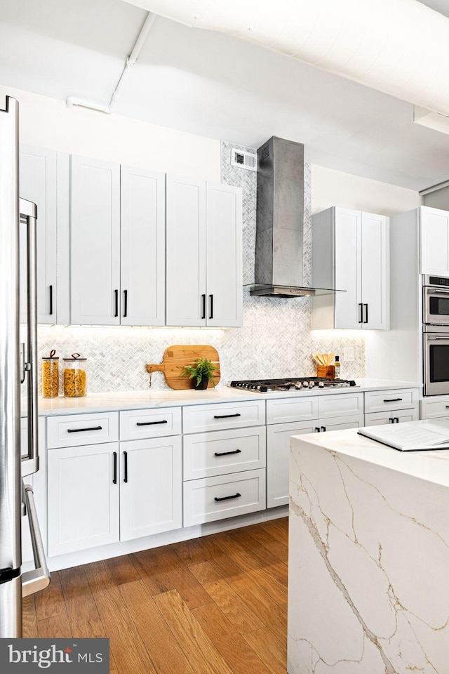 kitchen featuring appliances with stainless steel finishes, wall chimney exhaust hood, white cabinets, light stone counters, and hardwood / wood-style floors