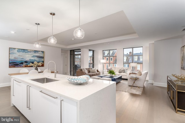 kitchen with a sink, light wood-style floors, hanging light fixtures, an island with sink, and a raised ceiling