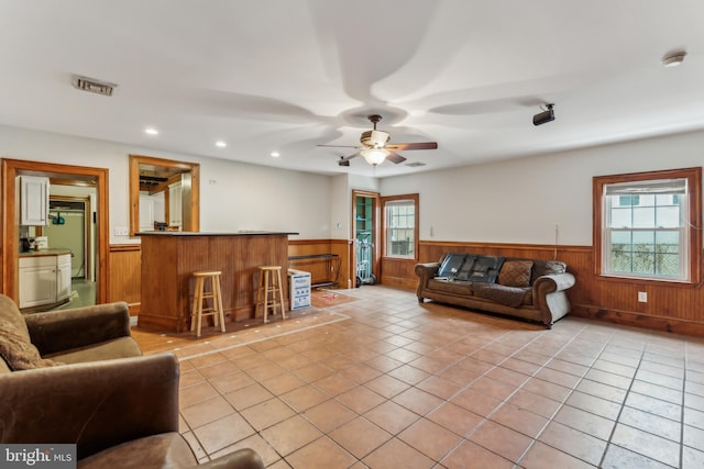 living room with light tile patterned floors, wooden walls, ceiling fan, and indoor bar