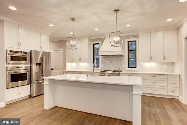 kitchen featuring a center island with sink, custom exhaust hood, light countertops, appliances with stainless steel finishes, and white cabinetry