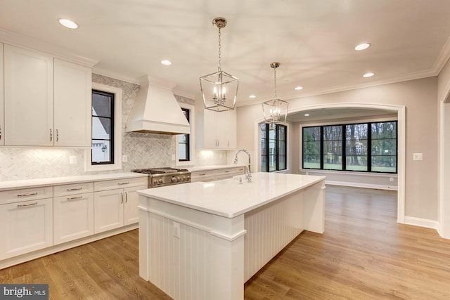 kitchen with a center island with sink, custom range hood, decorative light fixtures, stainless steel stove, and white cabinetry