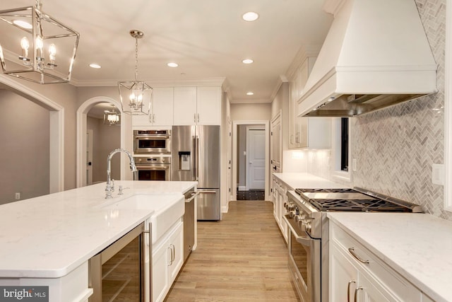 kitchen with a kitchen island with sink, premium range hood, white cabinetry, hanging light fixtures, and appliances with stainless steel finishes