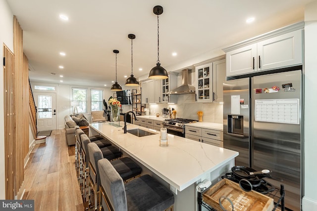 kitchen featuring a large island with sink, wall chimney range hood, stainless steel appliances, sink, and hanging light fixtures