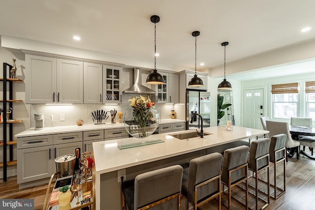 kitchen featuring an island with sink, dark wood-type flooring, pendant lighting, wall chimney exhaust hood, and sink