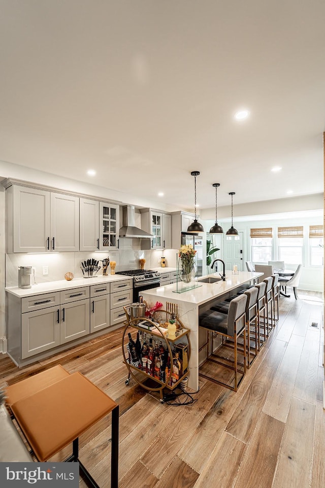 kitchen featuring an island with sink, gas stove, hanging light fixtures, wall chimney exhaust hood, and light hardwood / wood-style flooring