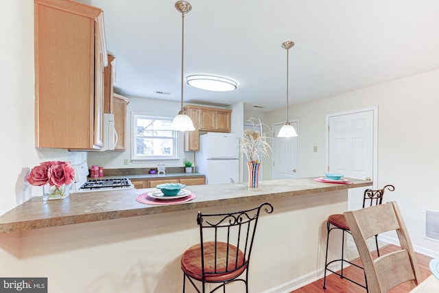 kitchen with kitchen peninsula, white appliances, a kitchen breakfast bar, light brown cabinetry, and hanging light fixtures