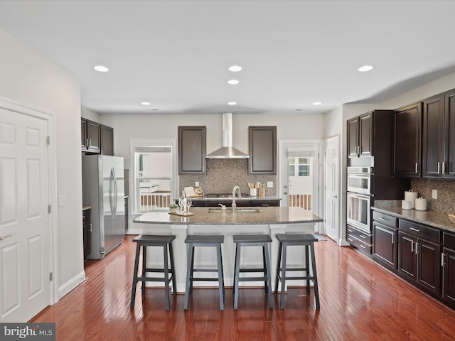 kitchen with wall chimney range hood, a kitchen island with sink, a breakfast bar area, and stainless steel fridge