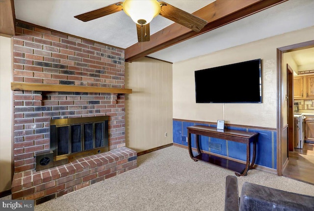 carpeted living room with a brick fireplace, beamed ceiling, and ceiling fan