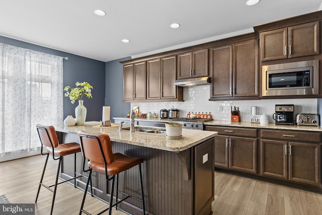 kitchen with sink, a kitchen island with sink, stainless steel microwave, and light stone counters