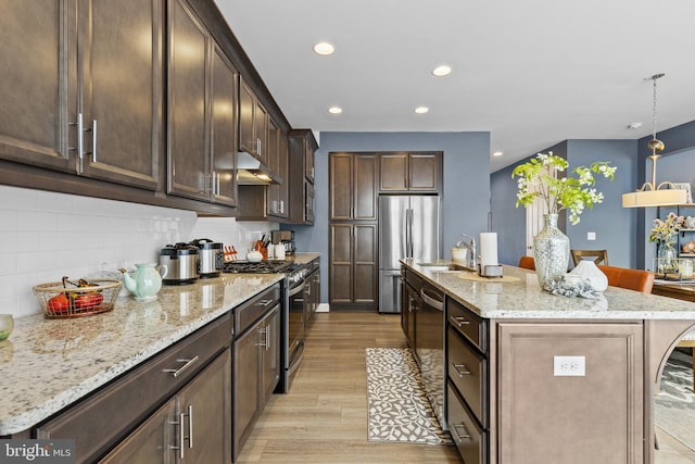 kitchen with hanging light fixtures, light wood-type flooring, an island with sink, stainless steel appliances, and light stone countertops