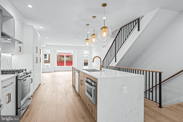 kitchen with white cabinets, wall chimney exhaust hood, stainless steel appliances, hanging light fixtures, and a center island with sink