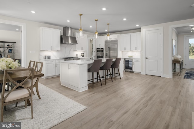 kitchen with wine cooler, decorative light fixtures, a center island with sink, wall chimney range hood, and white cabinets