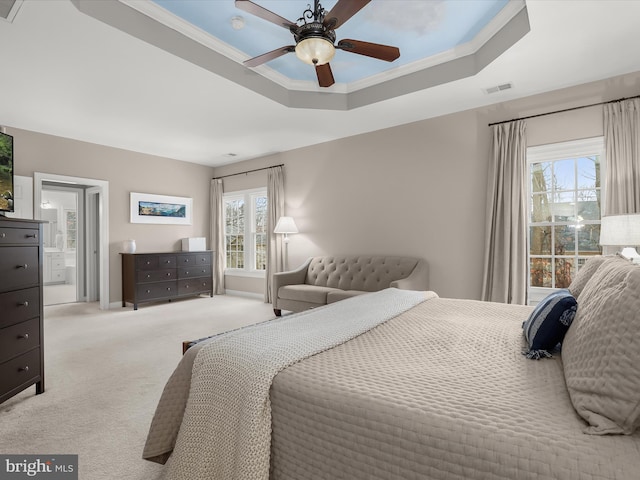 bedroom with a tray ceiling, crown molding, light colored carpet, visible vents, and ensuite bath