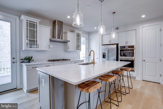 kitchen featuring a kitchen island with sink, wall chimney range hood, white cabinetry, and appliances with stainless steel finishes